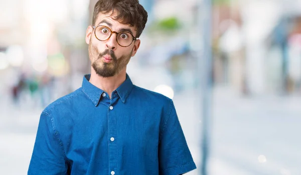 Young handsome man wearing glasses over isolated background making fish face with lips, crazy and comical gesture. Funny expression.
