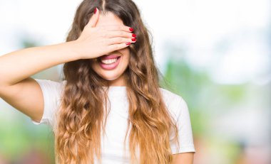 Gülümsüyor ve gözleri için sürpriz kapsayan yüz el ile gülüyor casual beyaz t-shirt giyen genç güzel kadın. Kör kavramı.