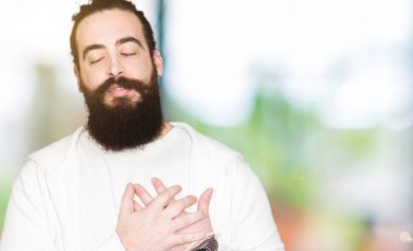 Uzun saç ve sakal ile sportif kazak gülümseyen giyen genç adamla kapalı gözler ve yüzünde minnettar jest ile göğüs üzerinde eller. Sağlık kavramı.