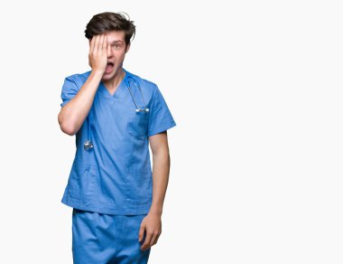 Yüz ve sürpriz duygu kendine güvenen gülümseme ile el ile bir göz kaplayan izole arka plan üzerinde tıbbi üniforma giyen genç doktor.