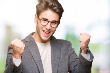 İzole arka plan üzerinde kutluyor gözlük takan genç iş adamı şaşırttı ve başarı için kaldırdı kollarını açarak hayret ve gözleri açık. Kazanan kavramı.