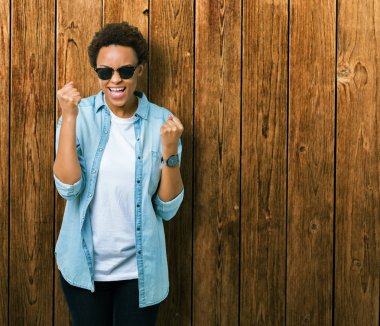 Güzel genç Afro-Amerikan kadın üzerinde güneş gözlüğü takıyor arka plan çok mutlu ve heyecanlı kaldırdı, silah ile kazanan jest yapıyor gülümseyen ve başarı için çığlık izole. Kutlama kavramı.