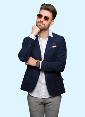 Çene düşünme hakkında soru, dalgın ifade üzerine güneş gözlüğü ve el ile moda ceket giyen genç yakışıklı zarif adam. Düşünceli yüzüyle gülümsüyor. Şüphe kavramı.