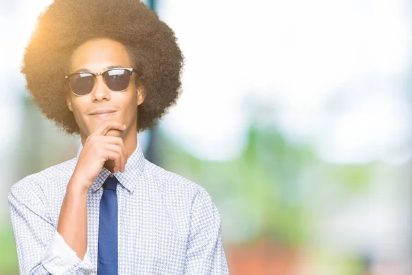 アフロ髪に関する質問は 物思いにふける式あご思考に手でサングラスをかけている若いアフリカ系アメリカ人ビジネスマン思いやりのある顔をして笑っています 疑いのコンセプト — ストック写真