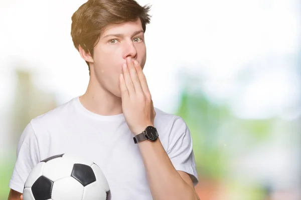 Young man holding soccer football ball over isolated background cover mouth with hand shocked with shame for mistake, expression of fear, scared in silence, secret concept