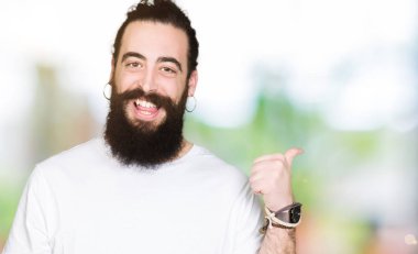 Uzun saçlı ve sakal ile genç hipster adam rahat beyaz t-shirt mutlu yüz görünümlü ve başparmak yukarı ile yan işaret gülümseyen giyiyor.