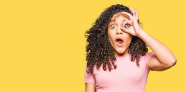 Pembe tişört giyen, kıvırcık saçlı genç bir kadın. Şok olmuş, şaşırmış bir yüz ve parmaklarından bakan bir göz hareketi yapıyor. İnançsız ifade.