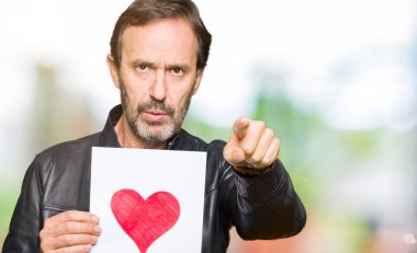 Yakışıklı orta yaş erkek holding kağıt kırmızı kalp kamera ve size parmak ile işaret ile el işareti, pozitif ve kendinden emin bir jest ön