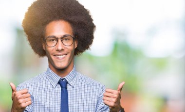 El ile pozitif jest yapıyor gözlük başarı işareti giyiyor afro saçlı genç Afro-Amerikan iş adamı başparmak gülümseyen ve mutlu. Neşeli bir ifadeyle, kazanan jest kameraya bakıyor.