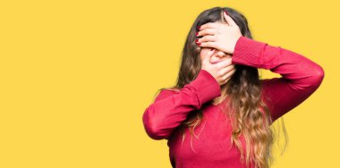 Kırmızı kazak giyen genç güzel kadın gözleri ve ağzı elleriyle kapamış, şaşırmış ve şok edir. Duyguyu gizleme