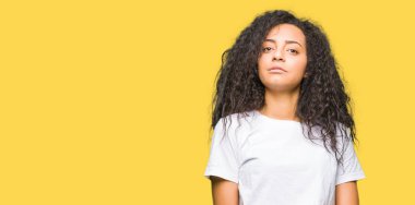 Genç güzel kız casual beyaz t-shirt eller birlikte giyen kıvırcık saçları ve parmak ile rahat ve neşeli gülümseyen geçti. Başarı ve iyimser