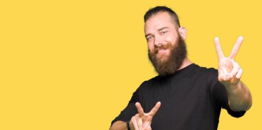 Genç Sarışın giyiyor rahat t-shirt gülümseyen kamera zafer işareti yapıyor parmak gösteren arayan adam. İki numaralı.