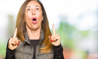 Kış yelek giyen güzel orta yaş kadın hayrete ve yukarıya bakıyor ve parmakları ile işaret ve silah kaldırdı şaşırttı.