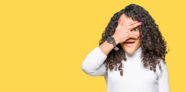 Kıvırcık saçlı, balıkçı yaka kazak giyen genç ve güzel bir kadın şok içinde yüzünü ve gözlerini kapayarak utangaç bir ifadeyle parmaklarının arasından bakıyor..