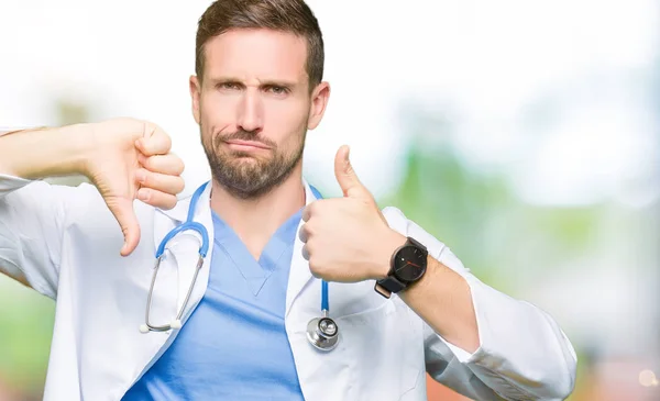 Handsome doctor man wearing medical uniform over isolated background Doing thumbs up and down, disagreement and agreement expression. Crazy conflict