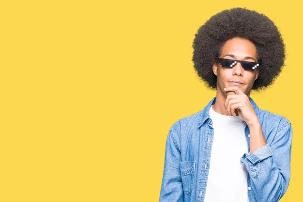 Young african american man with afro hair wearing thug life glasses with hand on chin thinking about question, pensive expression. Smiling with thoughtful face. Doubt concept.