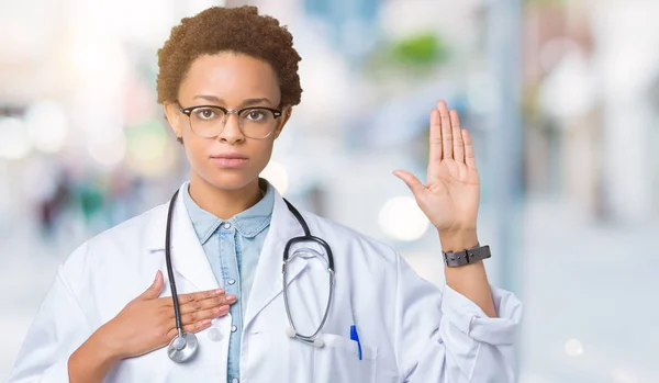 Young african american doctor woman wearing medical coat over isolated background Swearing with hand on chest and open palm, making a loyalty promise oath