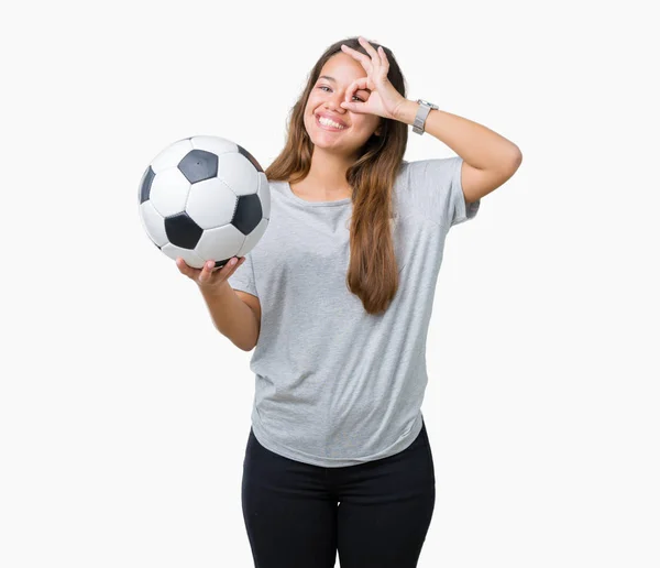 年轻的美丽的黑发女子拿着足球在孤立的背景与愉快的面孔微笑做确定的标志用手在眼睛看通过手指 — 图库照片