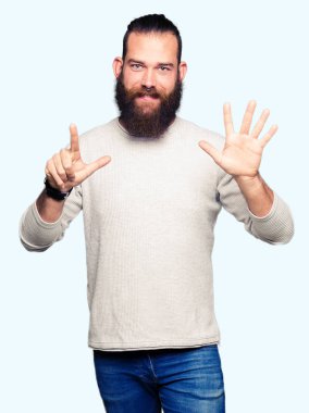 Genç sarışın adam rahat kazak gösteren ve parmak sayısı yedi ile işaret giymiş iken emin ve mutlu gülümseyen.