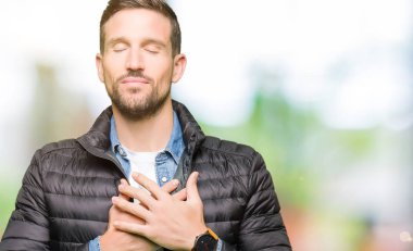 Yakışıklı adam ile gülümseyen kış ceket giyen kapalı gözler ve yüzünde minnettar jest ile göğüs üzerinde eller. Sağlık kavramı.