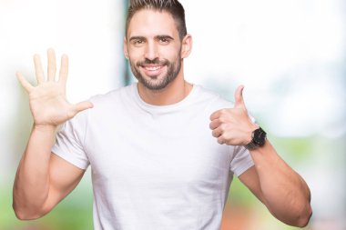 Gösterilen ve parmakları ile işaret izole arka plan üzerinde rahat beyaz t-shirt giyen genç adam kendinden emin ve mutlu gülümseyerek süre 6 numara.