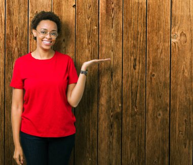 Güzel genç Afro-Amerikan kadın neşeli sunulması ve kameraya bakarak avucunun ile işaret gülümseyen izole arka plan üzerinde gözlük.