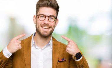 Kendinden emin gösterilen ve işaret parmakları diş ve ağız ile gülümseyen gözlüklü genç yakışıklı iş adamı. Sağlık kavramı.