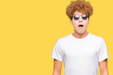 Afro saçlı Genç yakışıklı adam güneş gözlüğü korkuyor ve sürpriz ifade, korku ve heyecanlı yüz ile şok giyiyor.