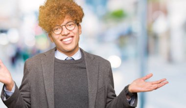 Genç yakışıklı iş adamı ile gözlük clueless ve şaşkın bir ifadeyle kollar ve eller kaldırdı giyen afro. Şüphe kavramı.