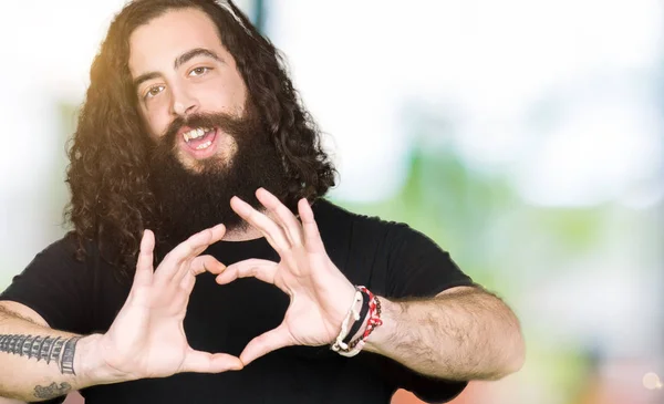 Young man with long hair and beard wearing heavy metal black outfit smiling in love showing heart symbol and shape with hands. Romantic concept.