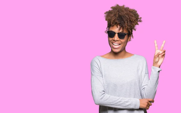 美丽的年轻非洲裔美国妇女戴着太阳镜在孤立的背景微笑着与幸福的脸眨眼在相机做胜利的标志 — 图库照片