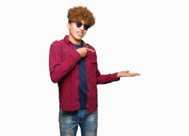 Hayret ve el ile sunulması ve parmak ile işaret ederken kameraya gülümseyen güneş gözlüğü takıyor afro saçlı Genç yakışıklı adam.