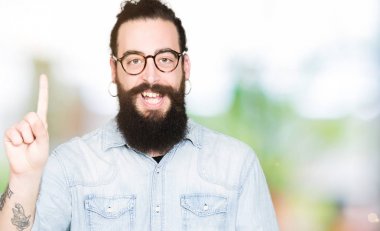 Uzun saç ve sakal gösterilen ve kendinden emin ve mutlu gülümseyen bir parmak ile işaret eden oktan yukarı gözlüklü genç hipster adamla.