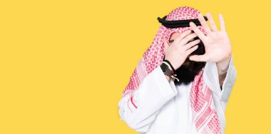 Geleneksel keffiyeh giymiş uzun saçlı Arap iş adamı elleriyle kapsayan gözleri eşarp ve stop jest ile acı ve korku ifadesi. Utandım ve negatif kavramı.