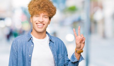 Afro saçlı Genç yakışıklı adam kot ceket giymiş mutlu yüz zafer işareti yapıyor kamera göz kırpıyor ile gülümsüyor. İki numara.