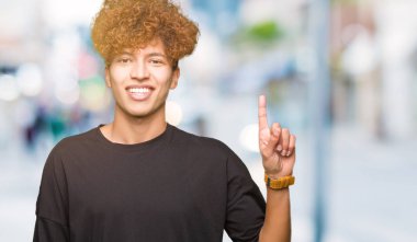 Genç yakışıklı adam ile afro saç giyen siyah t-shirt gösteren ve işaret yukarıya ile parmak sayı bir süre gülümseyen emin ve mutlu.