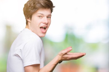 Yakışıklı delikanlı üzerinde rahat beyaz t-shirt giyen izole arka plan el ve reklam mutlu ve kendinden emin gülümseyen sunulması avuç, yanında işaret