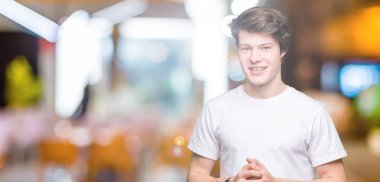 Arka plan eller üzerinde rahat beyaz t-shirt giyen genç yakışıklı adam birlikte izole ve rahat ve neşeli gülümseyen parmak çarpı işareti. Başarı ve iyimser