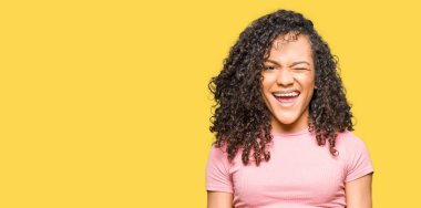 Kıvırcık saçlı pembe tişört göz kırpıyor seksi bir ifadeyle, neşeli ve mutlu yüzünü kameraya bakarak giyen genç güzel kadın.