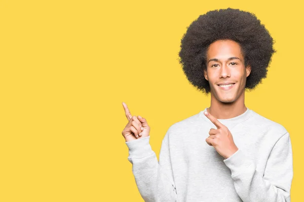 年轻的非洲裔美国男子 头发与运动运动衫微笑 看着相机指着两只手和手指的一侧 — 图库照片