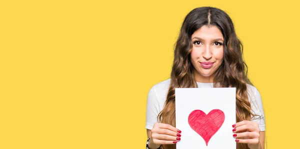 Young adult woman holding card with red heart with a confident expression on smart face thinking serious