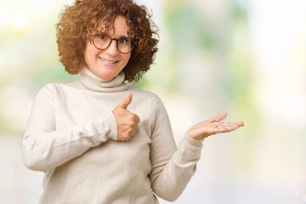 分離背景表示パーム手にタートルネックのセーターと眼鏡を着て 幸せで陽気な笑みを浮かべて親指で のジェスチャーをしている年配の女性を美しい中間エイガー — ストック写真