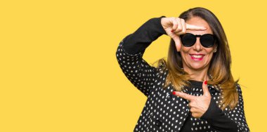 Güzel orta yaş kadın elle yapma çerçeve ve parmakları ile mutlu yüz gülümseyen güneş gözlüğü takıyor. Yaratıcılık ve Fotoğrafçılık kavramı.