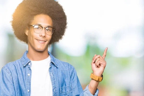 Young african american man with afro hair wearing glasses with a big smile on face, pointing with hand and finger to the side looking at the camera.