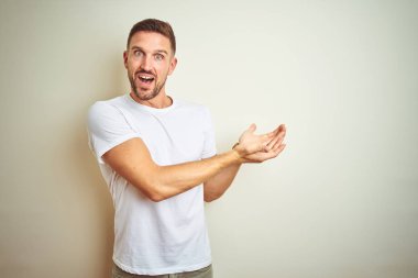 İzole edilmiş arka plan üzerine gündelik beyaz tişört giyen genç yakışıklı bir adam avuç içleri açık, fotokopi alanını gösteren, gülümseyerek mutlu bir şekilde gülümseyen bir reklam sunuyor.