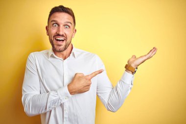 Sarı izole edilmiş arka plan üzerinde zarif beyaz gömlek giyen genç yakışıklı adam hayrete düşmüş ve kameraya gülümserken eliyle ve parmağıyla işaret ediyordu..