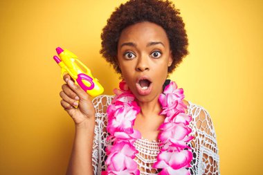 Afro saç giyen çiçek Hawaii Lei ve su tabancası ile genç Afrikalı Amerikalı kadın sürpriz bir yüz ile şok korkuyor ve korku ifadesi ile heyecanlı korkmuş