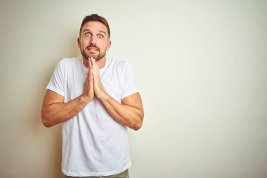 Ellerle birlikte AF emin gülümseyen için soran dua izole arka plan üzerinde rahat beyaz t-shirt giyen genç yakışıklı adam.