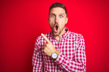 Kırmızı izole edilmiş arka plan üzerinde genç yakışıklı bir adam. Parmağını yana doğru göstererek şaşırmış, ağzı açık, şaşkın bir ifade..