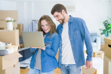 Yeni bir daireye taşımak için mutlu karton kutular etrafında bir odada bilgisayar laptop ayakta kullanarak genç çift